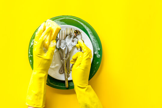 Washing dishes. Hands in rubber gloves and plates on yellow background top view cope space