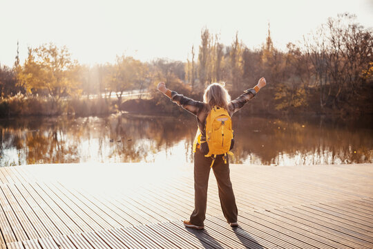 Happy woman standing with raised arms on wooden pier near forest lake