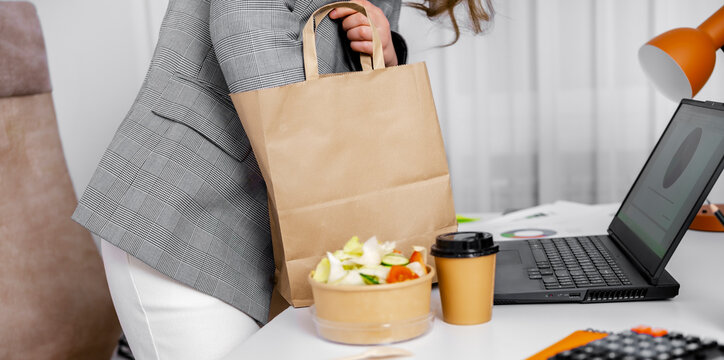 Business woman in suit holding paper shopping bag with lunch and coffee standing near desk in office manager executive corporate worker. Lunch break food delivery and busy lifestyle concept.