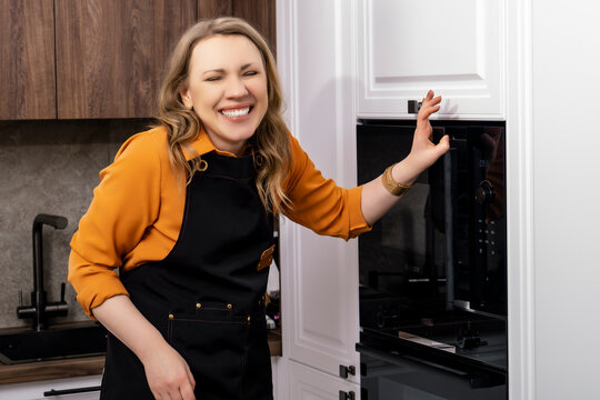 Cheerful young woman baker in apron laughs near black modern oven in stylish kitchen. Expression of sincere happiness and joy of domestic lifestyle.