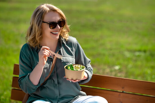 Happy woman in sunglasses eating green salad vegan poke bowl on park bench. Healthy diet nutrition and active wellness lifestyle outdoors.