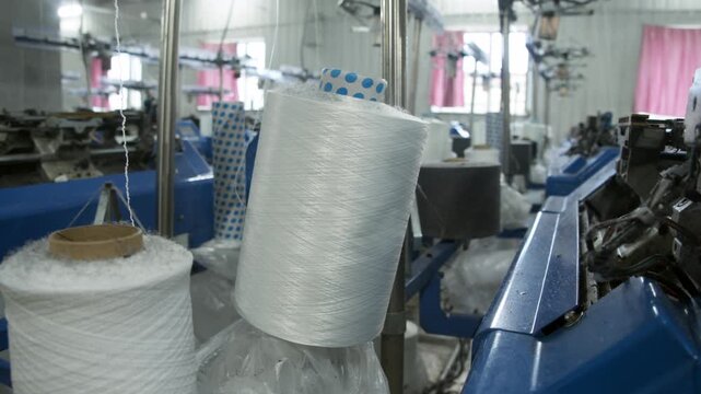 Industrial yarn spool delivers thread into textile knitting machine during manufacturing process. Factory equipment supports continuous fabric production.