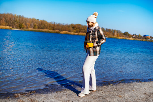 Woman walking by lake on sunny autumn day in beanie and warm plaid shirt holding thermos. Cold season walk and peaceful nature retreat.