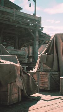 Wooden crates sit on a deck with cloth draped over them. The scene shows a rustic area with old structures. Sunlight shines down, highlighting the weathered wood.