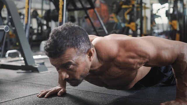 An Indian Asian fit, muscular, shirtless middle aged man performing a bodyweight resistance training exercise, push ups in a modern gym. Fitness, workout, healthy lifestyle, wellness, concept