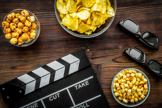 Crisp, popcorn, rusks for watching film. Clapperboard and glasses on dark wooden background top view