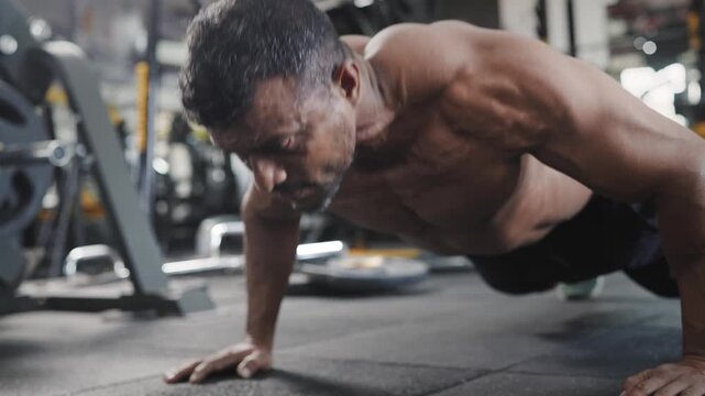 An Indian Asian fit, muscular, shirtless middle aged man performing a bodyweight resistance training exercise, push ups in a modern gym. Fitness, workout, healthy lifestyle, wellness concept