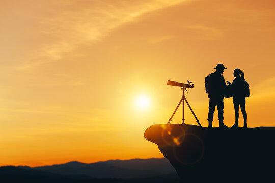 Silhouette of couple travelers with telescope and map on cliff at sunset. Concept of exploration, romantic journey, adventure, and future vision.