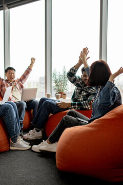 A diverse group of young adults celebrates a shared success while working together in a modern office space with beanbag chairs and laptops
