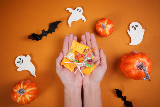 Hands holding assortment of Halloween candies over orange background with pumpkins and ghosts
