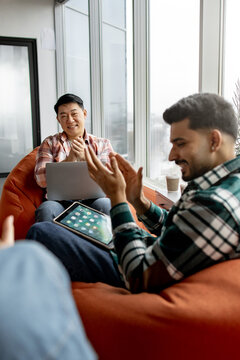 Two men in casual attire are sitting on beanbag chairs near a large window, one using a laptop and the other a tablet, both clapping enthusiastically