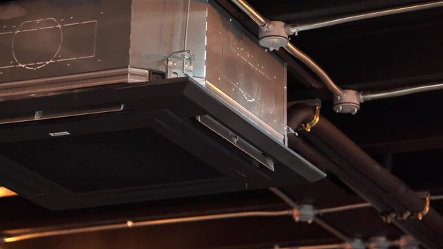 Black ceiling mounted cassette air conditioner installed on a dark industrial loft style ceiling with exposed pipes