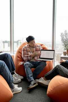 A man in a plaid shirt presents data on a laptop to colleagues sitting on beanbag chairs near a large window