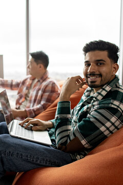 A smiling young man with a beard and dark hair is working on a laptop while sitting on an orange beanbag chair next to a window