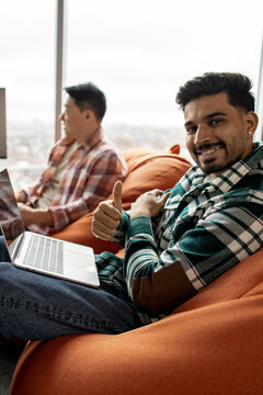 A young man with a beard smiles and gives a thumbs-up gesture while sitting with a laptop and another person in the background