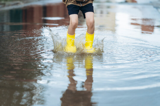 Boy in yellow rain boots splashes water in a summer puddle