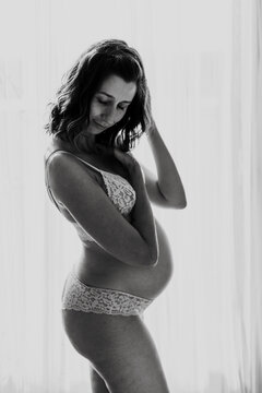 Pregnant woman in lace lingerie stands indoors looking at her belly