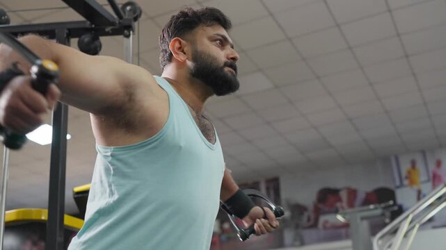 Low angle view of a young Indian Asian fit, muscular man performs a standing cable chest fly exercise during a resistance training session in a modern gym. Fitness, weight training workout concept