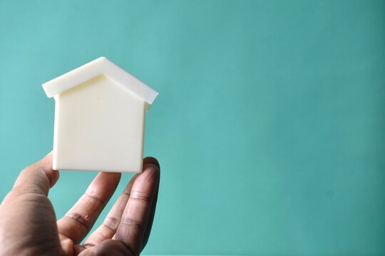 Close-up of a person's hand holding up a small white model house against a solid mint green studio background, representing real estate, property investment, or homeownership concepts.