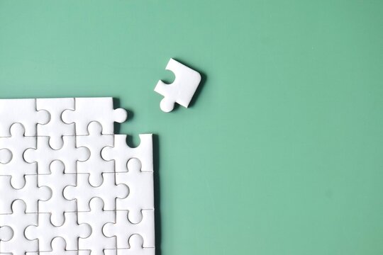 Flat lay top-down view of a partially assembled white jigsaw puzzle on a smooth sage green background, with a single detached piece lying near the vacant corner slot.