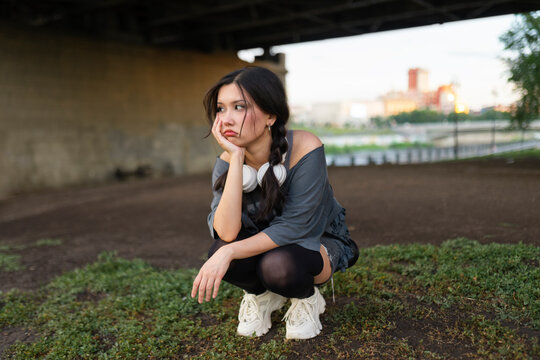 Bored woman with headphone squats under city bridge in summer