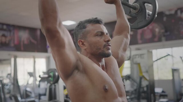 A young bare chested Indian Asian fit muscular male or man performs a barbell shoulder press during resistance weight training workout in modern gym. Fitness, healthy lifestyle, bodybuildig concept