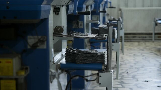 Finished construction glove exits automated machine and joins neatly stacked gloves on conveyor. Industrial equipment ensures continuous textile production with minimal manual handling.