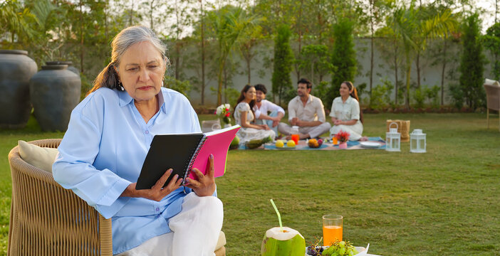 Indian old aged relax woman mom sitting on chair hold book read story novel outdoor house place happy male female man group talk speak spend quality time enjoy new trip day open in air garden lawn