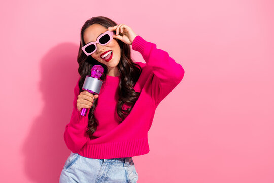 Young stylish woman sings into a pink microphone wearing a bright sweater against a pink background