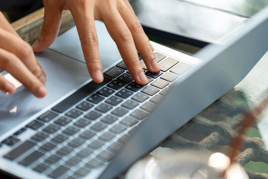 A close-up detail shot showing human hands typing on a modern silver laptop keyboard. Set in a bright, minimalist workspace or cafe environment, this image emphasizes concepts of remote work, software