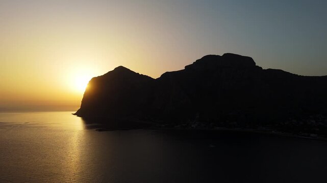 Dramatic sunrise silhouette of the Capo Gallo promontory in Palermo, Sicily. Aerial view showing the dark mountainous headland against dawn sky.