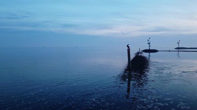 Drone slides above the Wadden Sea at Terschelling, Friesland, Netherlands, following navigation beacons, stone breakwaters and calm tidal water beneath the soft blue-hour sky.