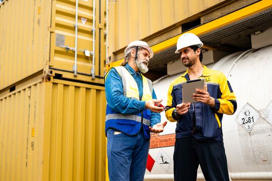 Two industrial workers in hard hats discuss logistics beside shipping containers and a tank, reviewing data on a tablet at a freight yard.