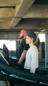 Two focused adults side by side on treadmills in a modern industrial gym, doing cardio training under soft daylight.