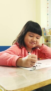 A young child in a cozy classroom draws with a marker on paper, focused and calm, resting her head as books and colorful decor sit in the background.