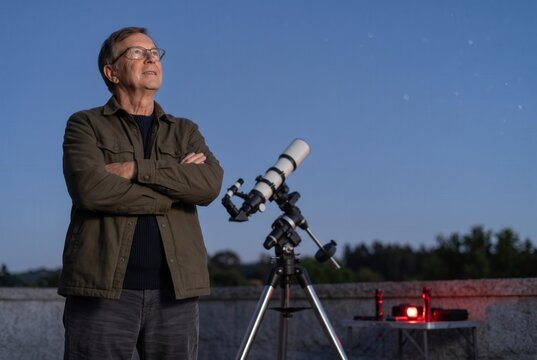 Mature man standing with arms crossed looking at the night sky next to a telescope