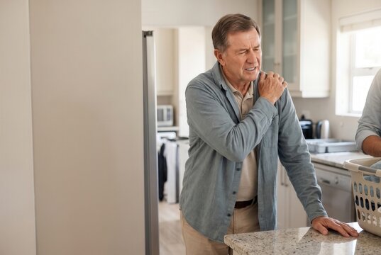 Senior man experiencing discomfort and shoulder pain, holding his injured joint in a home kitchen