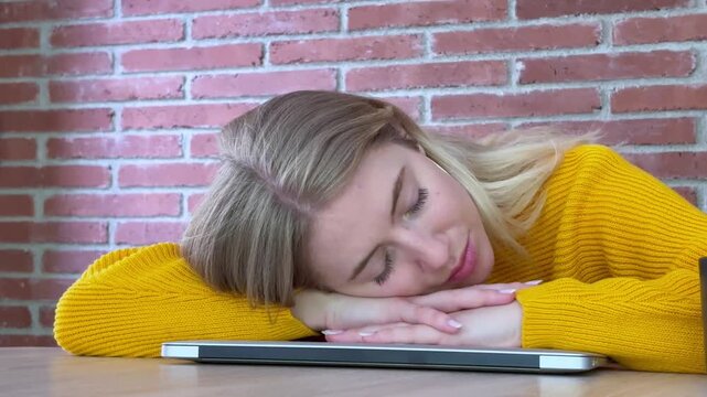 Exhausted young woman falling asleep on her closed laptop at a desk due to overworking, burnout, or fatigue, with a brick wall in the background representing a difficult study session