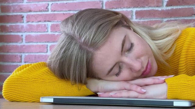 Overworked young woman falling asleep on her desk with her head resting on a closed laptop, exhausted from too much work or studying in a modern loft office with a brick wall background