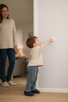 Toddler reaching up to press a light switch on a wall. Young boy turning on lights while a woman watches