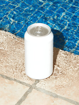 White soda can standing on edge of swimming pool near water on mosaic tiles.  Aluminum can with a carbonated beverage without a trademark.