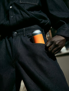 Orange-labeled soda can peeking out of pocket of man wearing dark jeans and dark shirt. Fizzy water in a standard tin can.