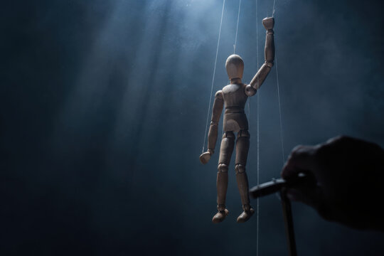 Wooden marionette puppet raised arm controlled by hand in spotlight with dark smoky background, symbolizing control and manipulation