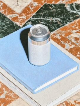 Aluminum soda can with white label standing on stack of books in room with multicolored tiled floor. Tin can with a liquid fizzy beverage.