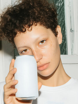 Curly-haired young woman in white t-shirt holding white aluminum chilled soda can against wall background. Fizzy soda water in a sealed metal packaging.