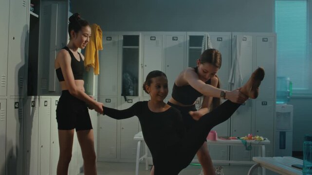 Teenage Female Athletes in Black Training Gear Helping Teammate Execute High Vertical Split Stretch inside Gym Locker Room. Concept of Professional Flexibility, Cooperation, and Group Warm Up Routine.