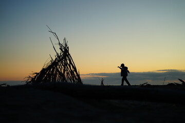 Silhouette of a child joyfully walking near a wooden teepee structure against a sunset sky. The scene captures a moment of playful exploration in a natural setting. © Nadezda Anokhina