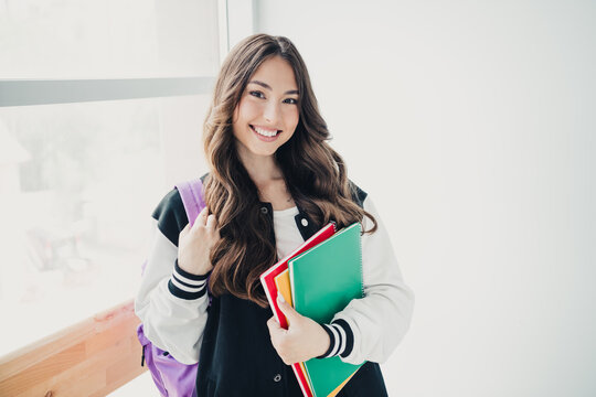 Smiling teenage student wearing a varsity jacket and backpack holding colorful notebooks in a bright school hallway