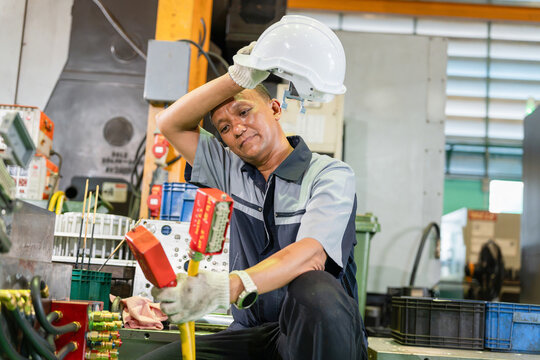 Tired industrial worker wiping sweat from forehead in manufacturing factory, Exhausted worker taking off hard hat and wiping sweat in industrial plant, Asian technician man resting after hard work