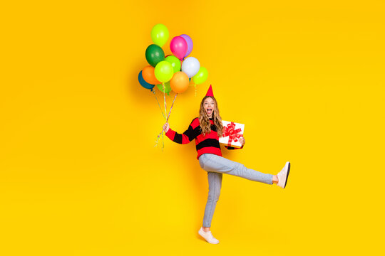 Cheerful woman in casual fashion holding balloons and a present on a vibrant yellow background for celebration
